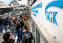 Bu görselde, Sosyal Güvenlik Kurumu (SGK) binasının önünde uzun bir kuyrukta bekleyen vatandaşlar yer alıyor. İnsanlar güvenlik bariyerlerinin arkasında sıralanmış ve bazıları ellerinde belgeler tutuyor. Ön planda, “ÖZEL GÜVENLİK” yazılı yelek giyen bir güvenlik görevlisi vatandaşlarla ilgileniyor. SGK binasının tabelası da sağ üst köşede net şekilde görülüyor.
