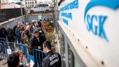 Bu görselde, Sosyal Güvenlik Kurumu (SGK) binasının önünde uzun bir kuyrukta bekleyen vatandaşlar yer alıyor. İnsanlar güvenlik bariyerlerinin arkasında sıralanmış ve bazıları ellerinde belgeler tutuyor. Ön planda, “ÖZEL GÜVENLİK” yazılı yelek giyen bir güvenlik görevlisi vatandaşlarla ilgileniyor. SGK binasının tabelası da sağ üst köşede net şekilde görülüyor.