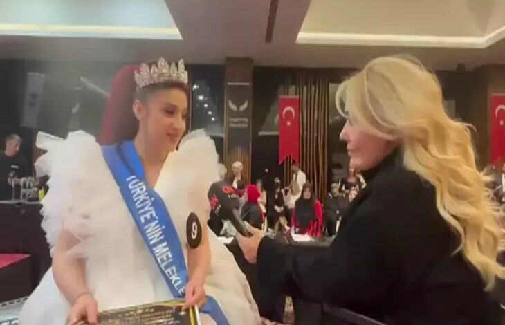 Fotoğrafta, tekerlekli sandalye kullanan iki kadın karşı karşıya duruyor. Sağ taraftaki kadının elinde bir mikrofon var. Sol taraftaki kadının ise üzerinde beyaz bir kıyafet ve “Meleklerin güzellik yarışması” yazısı yer alıyor.