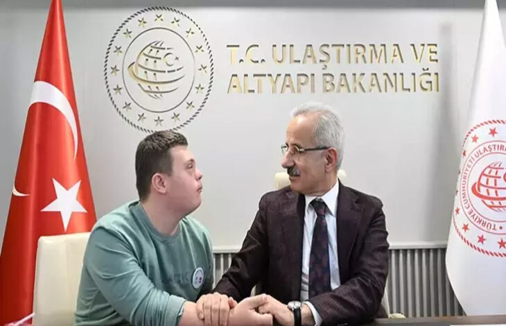 Ulaştırma ve Altyapı Bakanı Abdulkadir Uraloğlu, yanında yer alan Down sendromlu engelli birey ile bir masanın arkasında oturuyor ve konuşuyorlar. Bakanın arkasında ise Ulaştırma ve Altyapı Bakanlığı yazısı var.