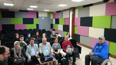 Bu görselde, modern ve renkli panellerle kaplı bir salonda bir grup insan oturuyor. Katılımcılar arasında kadınlar, erkekler, yaşlılar ve tekerlekli sandalyedeki bir birey de yer almakta. Ortam sıcak ve topluluk hissi veriyor. İnsanlar kameraya dönük şekilde gülümsüyor veya dikkatle bakıyor. Bu sahne, bir toplantı ya da sosyal etkinlik kapsamında bir araya gelmiş, çeşitli yaş ve özelliklere sahip bireyleri yansıtıyor.