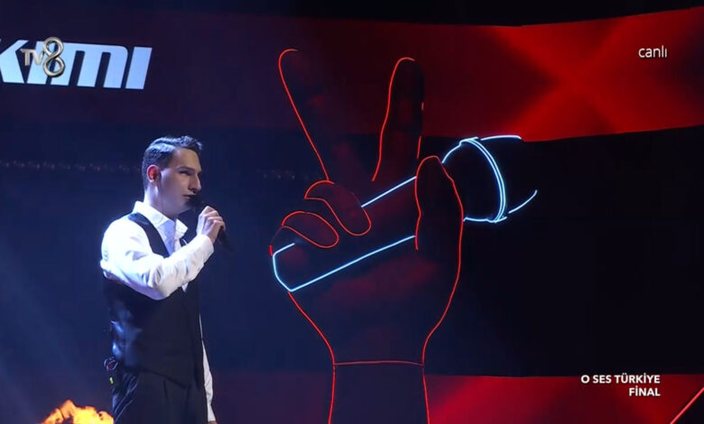 Ayakta elinde tuttuğu mikrofonla performans sergileyen Mert Özer'in yandan çekilen fotoğrafı.