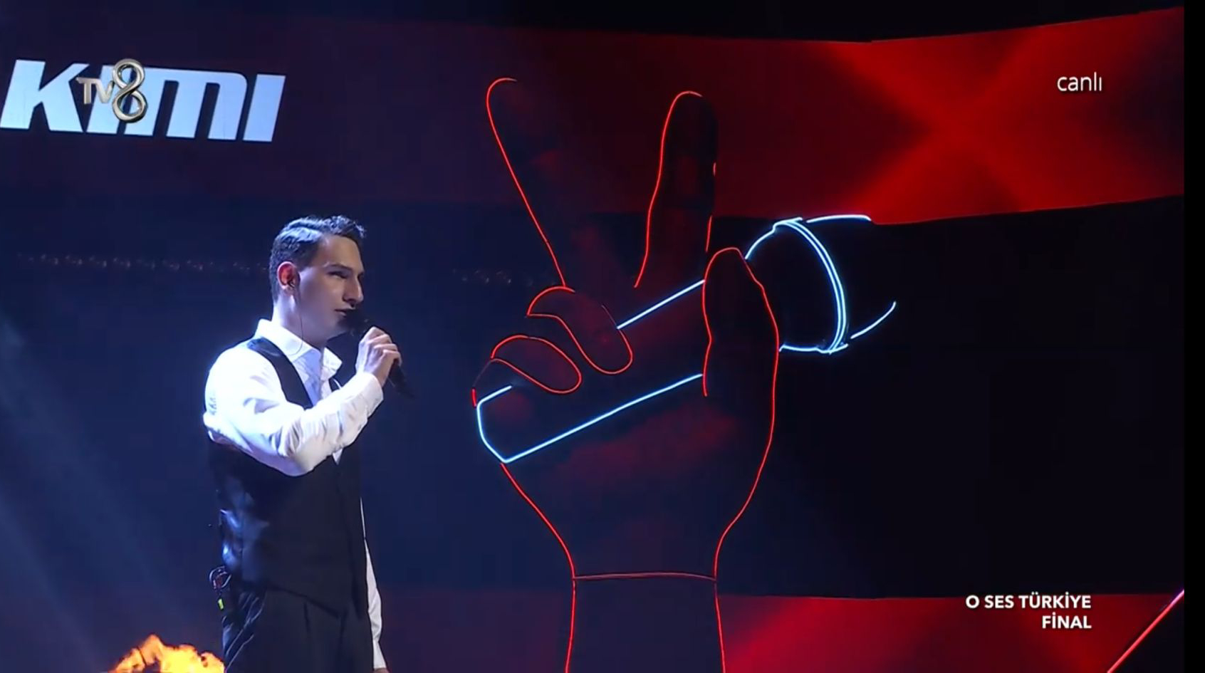 Ayakta elinde tuttuğu mikrofonla performans sergileyen Mert Özer'in yandan çekilen fotoğrafı.