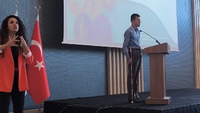 Görselde bir etkinlik sahnesi yer almakta. Sağda, görme engelli bir erkek bastonuyla kürsüde konuşma yapıyor. Mavi gömlek giymiş ve projeksiyon ekranına dönük. Sol tarafta ise kırmızı ceketli bir kadın işaret diliyle çeviri yapıyor. Arkada Türk bayrağı yer alıyor. Sahne üzerindeki sunum ekranında erişilebilirlikle ilgili simgeler görülüyor.