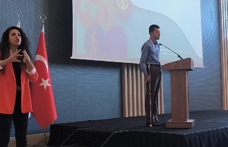 Görselde bir etkinlik sahnesi yer almakta. Sağda, görme engelli bir erkek bastonuyla kürsüde konuşma yapıyor. Mavi gömlek giymiş ve projeksiyon ekranına dönük. Sol tarafta ise kırmızı ceketli bir kadın işaret diliyle çeviri yapıyor. Arkada Türk bayrağı yer alıyor. Sahne üzerindeki sunum ekranında erişilebilirlikle ilgili simgeler görülüyor.