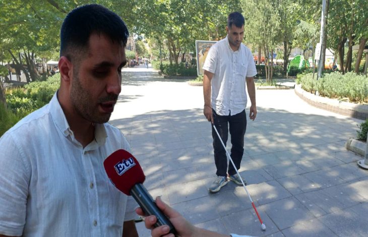 Görselde, açık havada ağaçlarla çevrili bir alanda iki erkek yer alıyor. Önde bulunan kişi mikrofon tutularak röportaj yapıyor; mikrofonun üzerinde "ETC" yazısı görülüyor. Arkada yer alan kişi ise beyaz baston kullanarak yürüyen doğuştan görme engelli bir birey. Her iki kişi de beyaz gömlek giymiş. Görsel, 6 Şubat depremi sonrası Manisa’ya taşınan ve iş arayan görme engelli Kamil Çelik’in kamuoyuna seslendiği bir anı yansıtıyor.