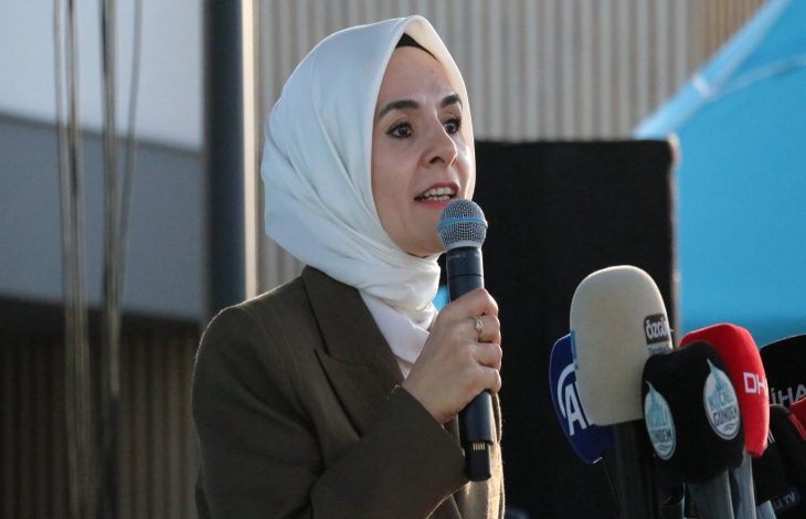 Görselde, başörtülü bir kadın açık havada mikrofonla konuşma yaparken görülüyor. Üzerinde koyu renk bir ceket var. Önünde çeşitli basın kuruluşlarına ait mikrofonlar yer alıyor (TRT, A Haber, DHA gibi). Arka planda mavi bir çadır ve modern bir bina detayı seçiliyor. Kadın ciddi ve etkileyici bir ifadeyle hitap ediyor.