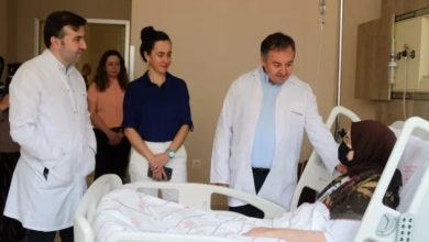 Görselde, hastane odasında bir hasta yatağında yatan başörtülü bir kadın yer alıyor. Kadının yanında beyaz önlük giymiş üç sağlık çalışanı bulunmakta; biri hastayla doğrudan konuşurken diğer ikisi ayakta durarak durumu izliyor. Arkada birkaç kişi daha görülüyor. Hasta, maskeli ve yorganla örtülmüş şekilde yatmakta. Odanın tıbbi donanımı ve serum seti de görselde yer alıyor.
