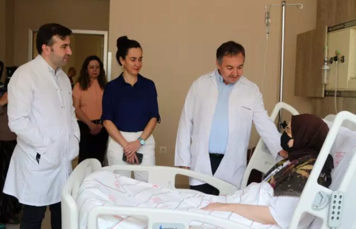 Görselde, hastane odasında bir hasta yatağında yatan başörtülü bir kadın yer alıyor. Kadının yanında beyaz önlük giymiş üç sağlık çalışanı bulunmakta; biri hastayla doğrudan konuşurken diğer ikisi ayakta durarak durumu izliyor. Arkada birkaç kişi daha görülüyor. Hasta, maskeli ve yorganla örtülmüş şekilde yatmakta. Odanın tıbbi donanımı ve serum seti de görselde yer alıyor.