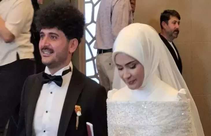 Görselde, gelinlik ve damatlık giymiş bir çift, mutlu bir şekilde yan yana duruyor. Damat papyonlu siyah bir takım elbise giymiş, gülümsüyor. Gelin ise beyaz başörtüsü ve ışıltılı bir gelinlik içinde, hafifçe başını eğmiş. Arka planda birkaç kişi daha görünüyor. Bu kare, bir düğün töreninden, sevgi ve mutluluğun paylaşıldığı özel bir anı yansıtıyor. Damat, aynı zamanda ampute futbolcu olarak biliniyor.