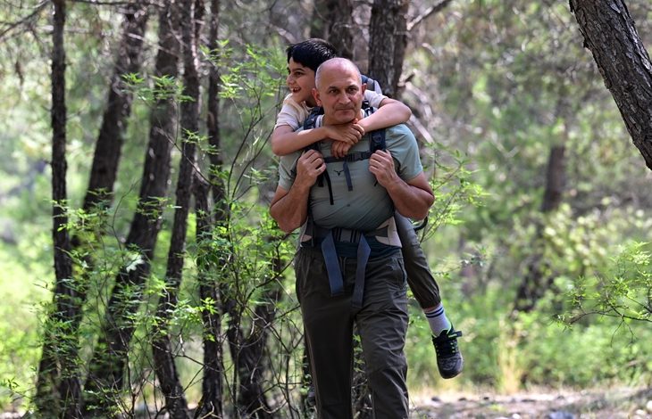 Görselde, ormanlık bir alanda yürüyen bir adamın sırtında bir çocuk taşıdığı görülüyor. Adam, çocuğu özel taşıma aparatına benzer bir düzenekle sırtına almış. Çocuk mutlu ve gülümsüyor. Doğa ile iç içe bu kare, engelli bir çocuğun babasıyla birlikte doğanın keyfini çıkardığı anlamlı ve duygu yüklü bir anı yansıtıyor.