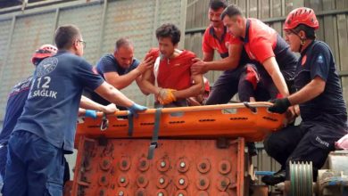 Görselde, bir grup kurtarma görevlisi ve sağlık personeli, ağır yaralanan bir işçiyi plastik kırım makinesinden sedyeyle çıkarmaya çalışıyor. Yaralı işçi kırmızı iş kıyafeti giymiş, oldukça acı çekiyor gibi görünüyor. Olay bir fabrikanın içinde geçiyor; arka planda sanayi tipi makineler ve metal yapı dikkat çekiyor. Kurtarma ekipleri organize bir şekilde çalışıyor, bazıları itfaiyeci, bazıları sağlık personeli.