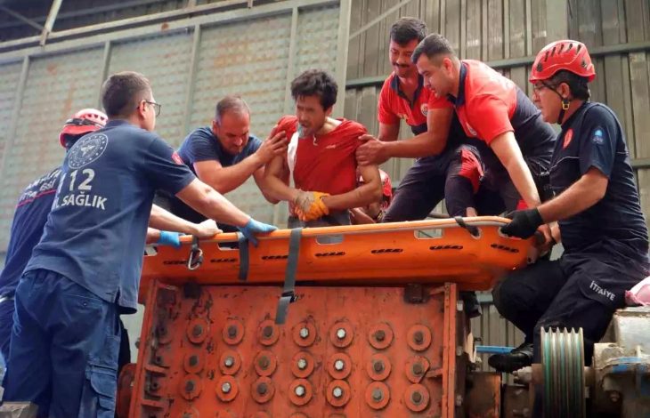 Görselde, bir grup kurtarma görevlisi ve sağlık personeli, ağır yaralanan bir işçiyi plastik kırım makinesinden sedyeyle çıkarmaya çalışıyor. Yaralı işçi kırmızı iş kıyafeti giymiş, oldukça acı çekiyor gibi görünüyor. Olay bir fabrikanın içinde geçiyor; arka planda sanayi tipi makineler ve metal yapı dikkat çekiyor. Kurtarma ekipleri organize bir şekilde çalışıyor, bazıları itfaiyeci, bazıları sağlık personeli.