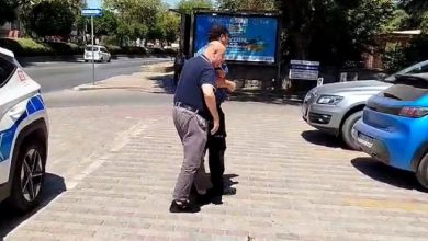 Görselde, Aydın’ın Efeler ilçesinde geçtiği anlaşılan bir olayda, iki görevli erkeğin görme engelli bir bireye yardım ettiği görülüyor. Kaldırımda yürüyen üç kişi bulunmakta; biri rehberlik eden görevli, diğeri görme engelli birey ve diğer kişi de destek sağlıyor. Arka planda park etmiş araçlar ve bir reklam panosu yer alıyor. Yardım edilen bireyin bankaya gitmek üzere çağrı merkezi aracılığıyla destek istediği anlaşılıyor.