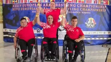 Görselde, dört sporcu tekerlekli sandalyede poz veriyor. Üzerlerinde kırmızı-siyah Türkiye formaları var. Arkalarında, “European Armwrestling Championship” yazılı mavi bir pankart bulunuyor. Sporculardan üçü ellerinde kupa tutuyor; bu, bir başarıyı simgeliyor. Arka planda çeşitli ülkelerin bayrakları yer almakta.