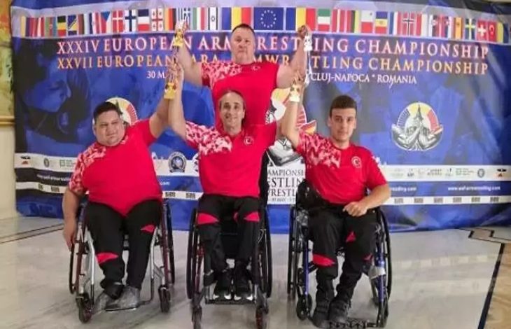 Görselde, dört sporcu tekerlekli sandalyede poz veriyor. Üzerlerinde kırmızı-siyah Türkiye formaları var. Arkalarında, “European Armwrestling Championship” yazılı mavi bir pankart bulunuyor. Sporculardan üçü ellerinde kupa tutuyor; bu, bir başarıyı simgeliyor. Arka planda çeşitli ülkelerin bayrakları yer almakta.