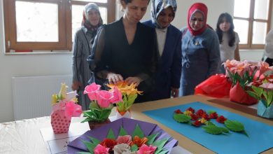 Görselde, bir masa üzerinde sergilenen renkli origami çiçekler yer alıyor. Ön planda, siyah elbise giymiş bir kadın elleriyle eserlerine dokunuyor. Arkasında ise başörtülü ve sivil giyimli birkaç kadın daha bulunuyor, dikkatle çalışmaları inceliyorlar. Gün ışığıyla aydınlanan bu oda, bir sergi alanı havası taşıyor.