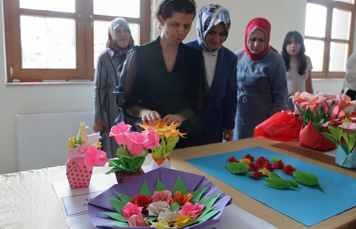 Görselde, bir masa üzerinde sergilenen renkli origami çiçekler yer alıyor. Ön planda, siyah elbise giymiş bir kadın elleriyle eserlerine dokunuyor. Arkasında ise başörtülü ve sivil giyimli birkaç kadın daha bulunuyor, dikkatle çalışmaları inceliyorlar. Gün ışığıyla aydınlanan bu oda, bir sergi alanı havası taşıyor.