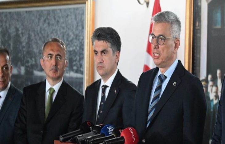 Görselde, resmi bir basın toplantısı ya da açıklama anı yer alıyor. Sağda yer alan kişi konuşma yaparken görülüyor ve önünde çeşitli basın kuruluşlarına ait mikrofonlar (TRT, DHA, A Haber vb.) bulunuyor. Sağlık Bakanı Kemal Memişoğlu’nun konuştuğu an olabilir. Diğer kişiler ciddi yüz ifadeleriyle onu dinliyor. Arka planda Türk bayrağı ve tarihi bir fotoğraf dikkat çekiyor.