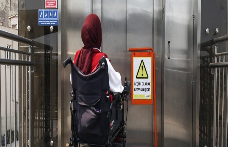 Görselde bir asansörün önünde duran akülü tekerlekli sandalye kullanan engelli birey yer alıyor. Asansörün önüne katılan tabelada geçici olarak hizmet dışı olduğu ifadesi belirtilmektedir.
