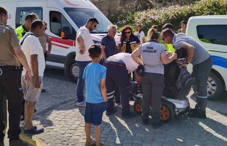 Fotoğrafta akülü tekerlekli sandalye üzerinde oturan engelli bireyin çevresinde sağlık personelleri ve bölgede bulunan insanlar yer alıyor. Görselde engelli birey ambulans ile hastaneye götürülmek için sedyeye taşınıyor.