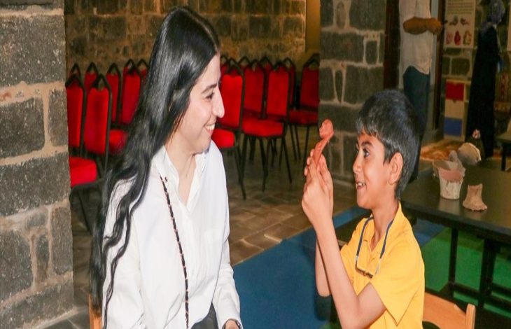 Fotoğrafta görme engelli bir çocuk kapalı bir ortamda tarihi bir eseri dokunarak anlamaya çalışıyor. Çocuğun yanında öğretmeni var.