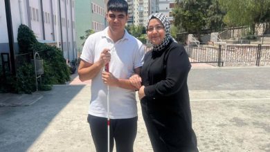 Görselde, açık bir alanda genç bir erkek ve bir kadın yan yana poz veriyor. Genç, beyaz baston tutuyor; bu, onun görme engelli olduğunu gösteriyor. Üzerinde beyaz bir tişört ve koyu renk pantolon var. Yanındaki kadın siyah kıyafetli ve başörtülü, gülümseyerek gence destek veriyor. Arka planda apartmanlar ve bir okul binası dikkat çekiyor.