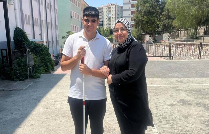 Görselde, açık bir alanda genç bir erkek ve bir kadın yan yana poz veriyor. Genç, beyaz baston tutuyor; bu, onun görme engelli olduğunu gösteriyor. Üzerinde beyaz bir tişört ve koyu renk pantolon var. Yanındaki kadın siyah kıyafetli ve başörtülü, gülümseyerek gence destek veriyor. Arka planda apartmanlar ve bir okul binası dikkat çekiyor.