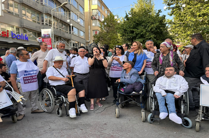 Bu görselde, Ankara’daki Sakarya Caddesi’nde gerçekleşen bir açık hava etkinliği görülüyor. Kalabalık bir grup, engelli bireylerin haklarına dikkat çekmek amacıyla toplanmış durumda. Katılımcıların arasında tekerlekli sandalye kullanan bireyler, yaşlılar ve çeşitli yaşlardan insanlar bulunuyor. Bazı kişilerin üzerindeki tişörtlerde “Engelliler seslerini duyurmak istiyor” ve “Haklarımızı istiyoruz” gibi ifadeler yer alıyor. Ortada mikrofonla konuşan bir kadın dikkat çekiyor.