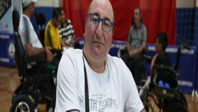 Görselde, spor salonu ortamında tekerlekli sandalye kullanan bir adam ön planda yer alıyor. Gözlüklü, beyaz tişört giymiş ve doğrudan kameraya bakıyor. Arka planda ise çeşitli yaş gruplarından yine tekerlekli sandalye kullanan bireyler oturuyor ve sohbet ediyor. Duvarda büyük bir Türk bayrağı asılı. Bu kare, engelli bireylerin dayanışma içinde bir araya geldiği sosyal ya da sportif bir etkinliği yansıtıyor.