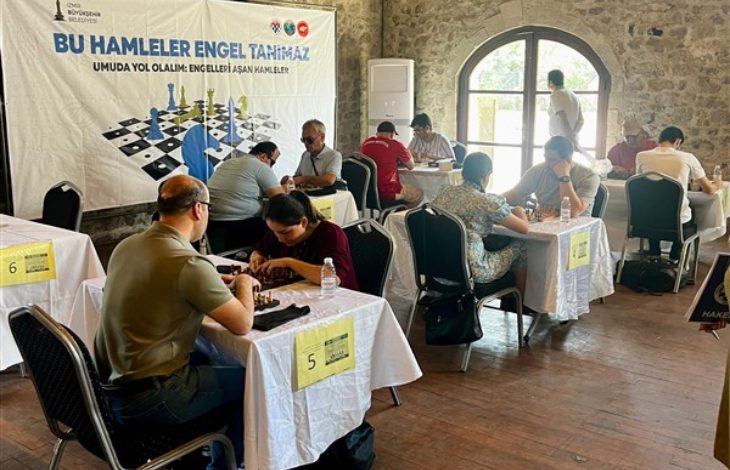 Görselde, taş duvarlı tarihi bir binada düzenlenen satranç turnuvası yer almakta. Masalarda karşılıklı oturan bireyler satranç oynuyor. Turnuvanın “Bu Hamleler Engel Tanımaz” sloganı dikkat çekiyor; afişte “Umuda yol olalım: Engelleri aşan hamleler” yazısı da yer almakta. Etkinlik, görme engelli bireylerin de katılım sağlayabildiği kapsayıcı bir organizasyon izlenimi veriyor. Masa numaraları ve su şişeleri, profesyonel bir turnuva ortamını yansıtıyor.