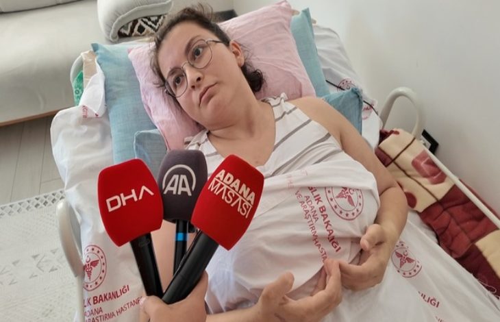 Görselde, hastane yatağında yatan genç bir kadın yer alıyor. Üzerinde Sağlık Bakanlığı’na ait hastane örtüsü var, yüzünde yorgun ama dirayetli bir ifade bulunuyor. Gözlük takıyor ve açık renkli bir atlet giymiş. Kadının önünde mikrofonlar tutulmuş – DHA, AA ve Adana Mavisi haber ajanslarına ait mikrofonlar net şekilde görülüyor.