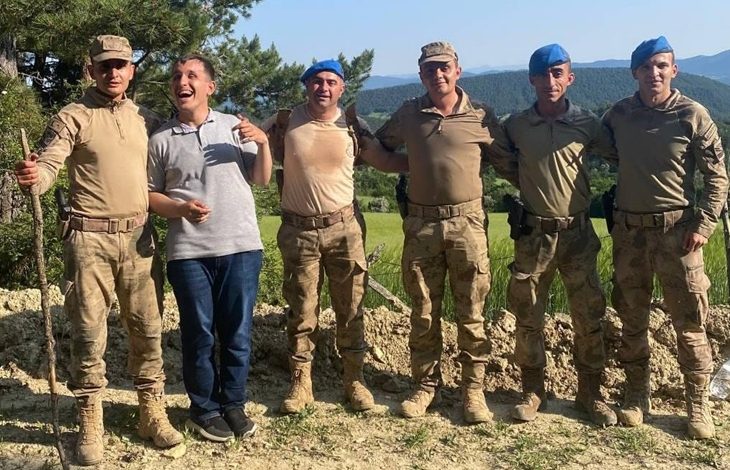 Bu fotoğrafta, ormanlık ve dağlık bir alanda, altı asker ve bir sivil erkek birlikte poz veriyor. Askerlerin çoğu kamuflaj üniformalı; üçü mavi bere takmış, diğerleri kasketli. Ortadaki sivil erkek gri tişört ve kot pantolon giymiş, neşeli ve gülümseyerek poz veriyor. Askerlerle samimi bir şekilde kol kola girmişler. Arka planda yeşillikler, ağaçlar ve uzak tepeler görünüyor.