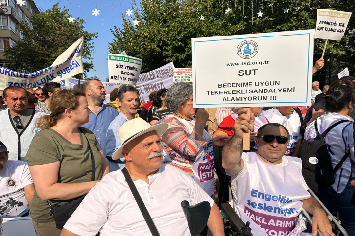 Bu görselde, engelli haklarına dikkat çekmek amacıyla düzenlenmiş bir protesto ya da farkındalık etkinliği yer almakta. Katılımcıların bir kısmı tekerlekli sandalye kullanıyor ve "Engelliler sesleniyor: Haklarımızı istiyoruz" yazılı tişörtler giymiş. Öndeki bir kişi “SUT – Bedenime uygun tekerlekli sandalyemi alamıyorum!” yazılı bir döviz taşıyor. Arka planda da benzer talepleri içeren başka dövizler görülüyor.