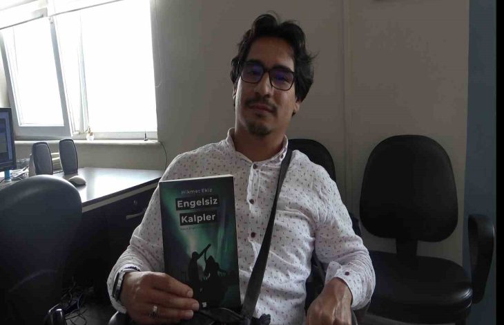 Görselde, gözlüklü bir erkek birey iç mekânda bir sandalyede oturuyor ve elinde “Engelsiz Kalpler” adlı kitabı tutuyor. Üzerinde beyaz, desenli bir gömlek var ve çapraz askılı bir çanta takıyor. Arka planda ofis ortamını andıran masa, bilgisayar ve boş sandalyeler görülüyor. Kitabın kapağında engelli bireylerin silüetleri yer almakta.