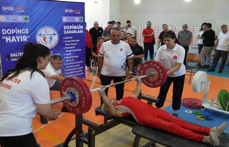 Görselde, Kırıkkale'de düzenlenen Bedensel Engelliler Halter Türkiye Şampiyonası’ndan bir an görülüyor. Kırmızı formasıyla bir sporcu bench press yaparken, etrafında antrenörler ve destek ekibi dikkatle bekliyor. Arkada “Dopinge Hayır” yazılı bilgilendirici pankartlar ve izleyiciler yer alıyor.