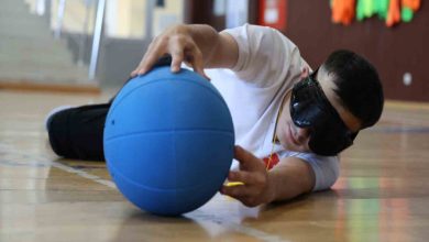 Gözleri siyah maskeyle kapalı erkek sporcu, mavi renkli golbol topunu iki eliyle tutup uzanıyor.