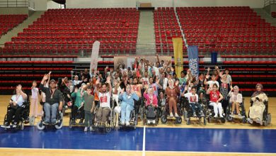 Görselde, büyük bir spor salonunun ortasında bir araya gelmiş tekerlekli sandalyedeki bireyler ve onlara eşlik eden kişiler yer alıyor. Grup, kameraya doğru neşeyle el sallıyor. Salonun tribünleri kırmızı koltuklarla dolu ama boş. Üst kısımda “Konya’yı Geçmişi Kadar Büyük, Görkemli Bir Geleceğe Hazırlıyoruz.” yazısı dikkat çekiyor. Arka planda etkinlik sponsorlarına ait bayraklar da yer almakta.