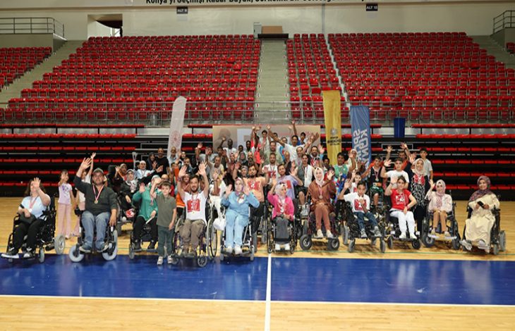 Görselde, büyük bir spor salonunun ortasında bir araya gelmiş tekerlekli sandalyedeki bireyler ve onlara eşlik eden kişiler yer alıyor. Grup, kameraya doğru neşeyle el sallıyor. Salonun tribünleri kırmızı koltuklarla dolu ama boş. Üst kısımda “Konya’yı Geçmişi Kadar Büyük, Görkemli Bir Geleceğe Hazırlıyoruz.” yazısı dikkat çekiyor. Arka planda etkinlik sponsorlarına ait bayraklar da yer almakta.