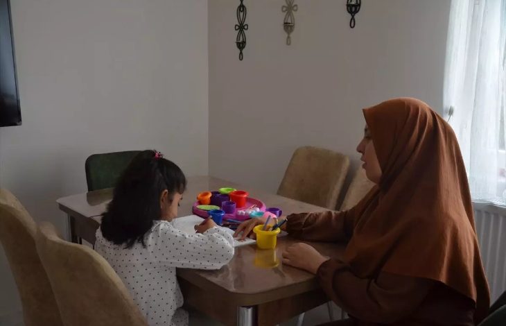 Fotoğrafta, orta yaşlarda olan bir kadın ve bir kız çocuğu masanın etrafında oturuyor. Çocuk önündeki kitaba boyama yapıyor ve masanın üzerinde farklı renklerde kutular var.