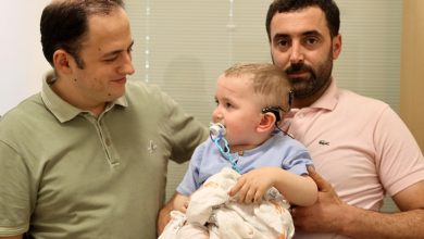 Görselde, iki yetişkin erkek arasında kucakta tutulan küçük bir çocuk yer alıyor. Çocuğun kulağında işitsel implant cihazı bulunuyor ve emzik emiyor. Yetişkinlerden biri çocuğa sevgiyle bakarken, diğeri kameraya doğru ciddi bir ifadeyle bakıyor. Sahne, çocuğun sağlıkla ilgili önemli bir anına işaret ediyor.