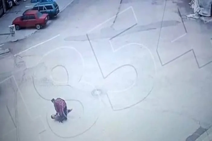 Bu görsel, bir güvenlik kamerası görüntüsünden alınmış ve oldukça sarsıcı bir anı yansıtıyor. Açık bir alanda, bir kişi yerde yatan başka bir kişiye saldırıyor gibi görünüyor. Saldırganın elinde silah kabzası olduğu belirtilmiş. Görselde, kırmızı ekoseli gömlek giyen şahıs net şekilde seçilebiliyor. Arka planda park etmiş araçlar ve boş bir cadde göze çarpıyor.