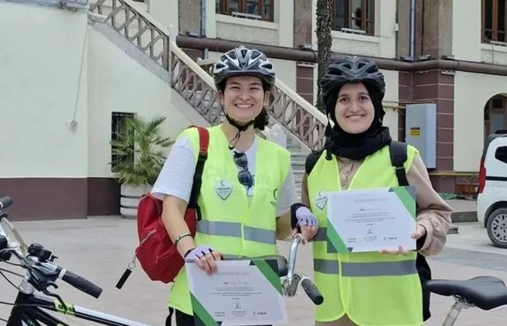 Görme engelli 2 kadın tandem bisiklet sürücüsü kadın birbirlerine açık yeşil renkte tulum benzeri bir kıyafet giymiş ve koruyucu kask takarak ayakta duruyorlar.