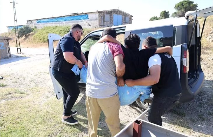 Görselde, dört kişi medikal ve ortopedik engeli bulunan bir kişiyi dikkatli bir şekilde araçtan indiriyor ya da araca bindiriyor. Engelli birey, mavi renkte bir taşıma örtüsüyle desteklenmiş. Arka planda kırsal bir alan ve eski yapılar görülüyor. Arazi düzensiz ama işlem dikkatli ve özenli bir şekilde yürütülüyor.