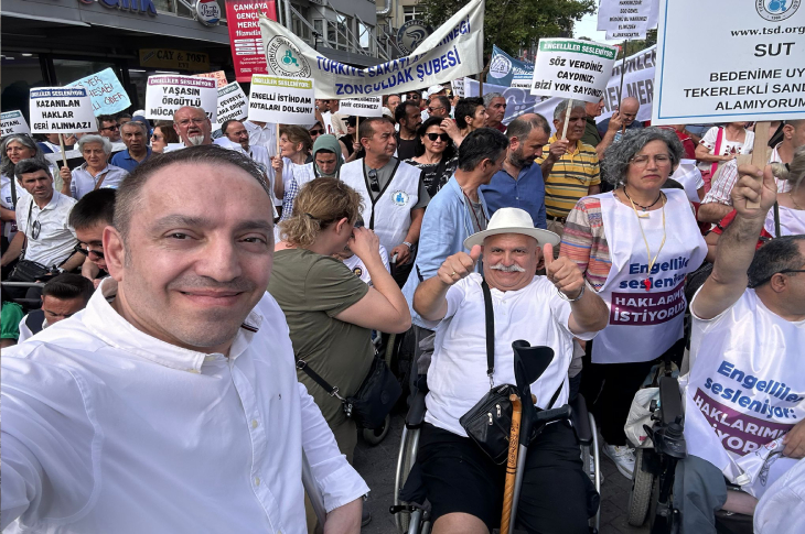 Bu görselde engelli bireyler ve destekçileri, hak taleplerini dile getirdikleri bir etkinlikte yer alıyor. Katılımcılar dövizler taşıyor; “Haklarımızı istiyoruz” ve “Tekerlekli sandalyemi alamıyorum” gibi mesajlar dikkat çekiyor. Ön planda Mehmet Kızıltaş özçekim yapıyor, etkinlik coşkulu ve kararlı bir atmosferde geçiyor.