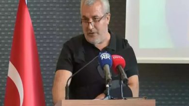 Görselde, bir erkek konuşmacı bir kürsüde basın mikrofonları önünde konuşma yapıyor. Adam siyah bir tişört giymiş, gözlük takıyor ve saçları gri. Kürsüde "AA" (Anadolu Ajansı) ve "DHA" (Demirören Haber Ajansı) mikrofonları bulunuyor. Arka planda Türkiye bayrağı ve projeksiyon ekranı yer alıyor. Ortam resmi ve profesyonel bir etkinlik atmosferi taşıyor.