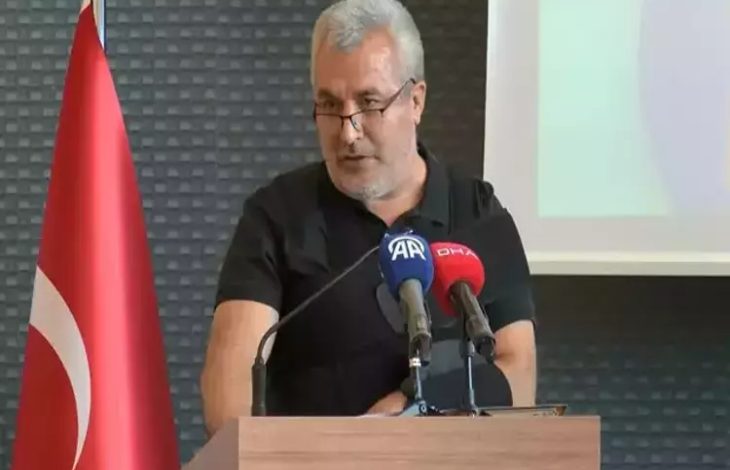 Görselde, bir erkek konuşmacı bir kürsüde basın mikrofonları önünde konuşma yapıyor. Adam siyah bir tişört giymiş, gözlük takıyor ve saçları gri. Kürsüde "AA" (Anadolu Ajansı) ve "DHA" (Demirören Haber Ajansı) mikrofonları bulunuyor. Arka planda Türkiye bayrağı ve projeksiyon ekranı yer alıyor. Ortam resmi ve profesyonel bir etkinlik atmosferi taşıyor.