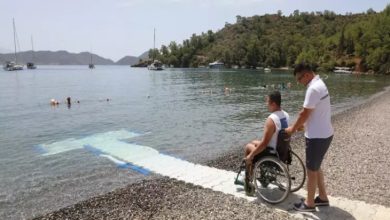 Görselde, bir engelli birey tekerlekli sandalyesiyle denize uzanan özel bir rampada, yanında ona eşlik eden bir kişiyle birlikte görülüyor. Arka planda sakin bir koy, yüzen insanlar ve demirlemiş tekneler var. Bu kare, Muğla’daki “Engelsiz Plajlar” projesi kapsamında engelli bireylerin denize erişimini kolaylaştırmak için yapılan düzenlemeleri yansıtıyor.