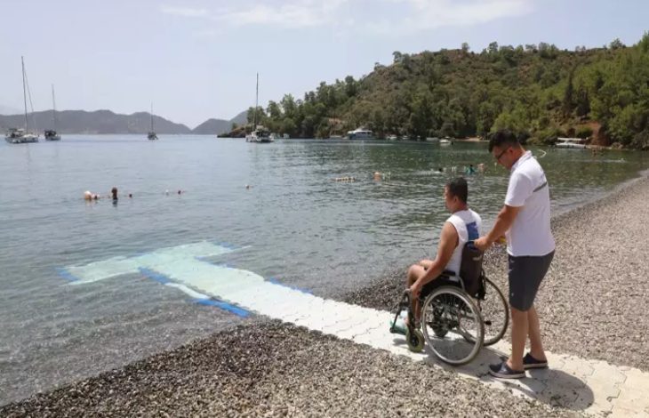Görselde, bir engelli birey tekerlekli sandalyesiyle denize uzanan özel bir rampada, yanında ona eşlik eden bir kişiyle birlikte görülüyor. Arka planda sakin bir koy, yüzen insanlar ve demirlemiş tekneler var. Bu kare, Muğla’daki “Engelsiz Plajlar” projesi kapsamında engelli bireylerin denize erişimini kolaylaştırmak için yapılan düzenlemeleri yansıtıyor.