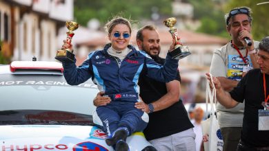 Görselde, yarış tulumu giymiş, güneş gözlüğü takan ve iki altın rengi kupa tutan gülümseyen bir kadın yer alıyor. Kadın, üzerinde bazı marka logoları (örneğin "HYPCO") bulunan beyaz bir ralli arabasının kaputunun üstünde oturuyor. Arkasında, sakallı bir erkek belinden onu tutmuş ve o da gülümsüyor. Sağ tarafta ise elinde mikrofon tutan başka bir adam konuşuyor gibi görünüyor. Arka planda birkaç kişi daha kısmen seçilebiliyor. Mekân açık hava gibi görünüyor; uzaklarda yeşillikler ve bazı binalar seçilebiliyor. Muhtemelen bir yarış etkinliği sonrası kutlama anı.